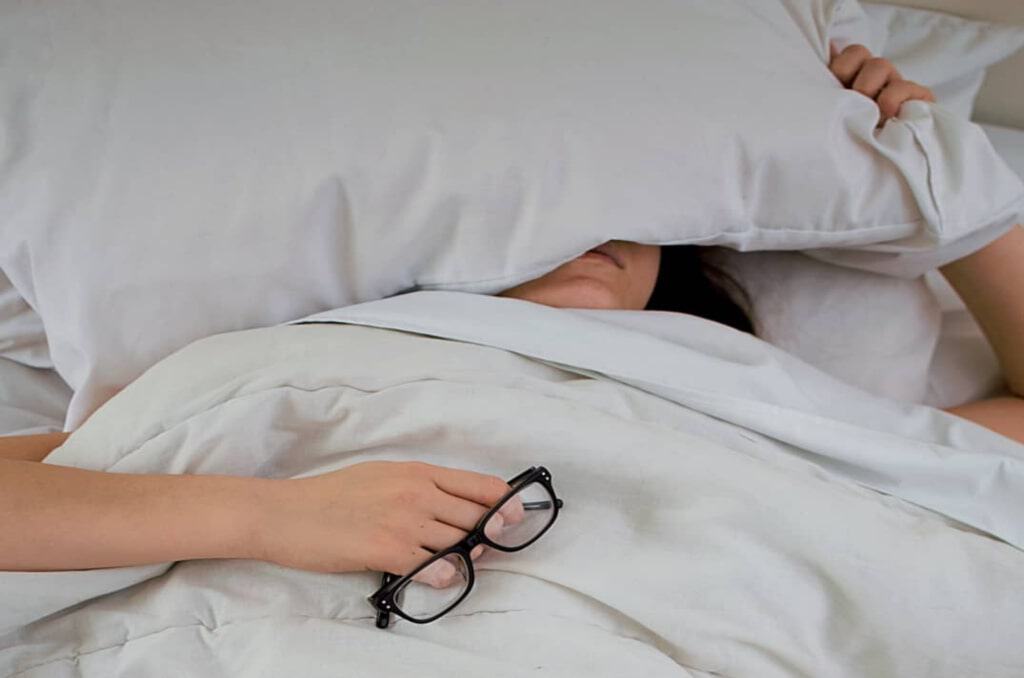 Woman lying in bed, holding glasses in one hand with the other resting on a pillow over her head, looking exhausted, symbolizing the importance of quality sleep.