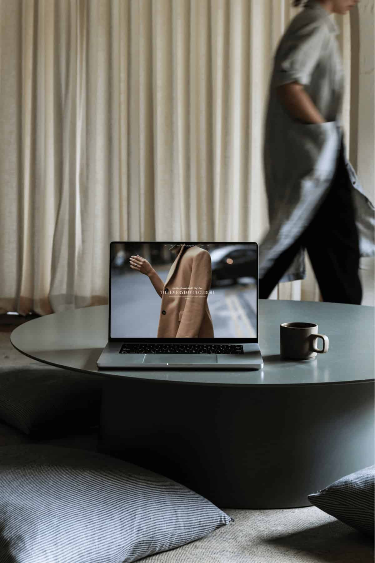 Laptop displaying The Everyday Flourish homepage on a glass-topped stone table, with a woman walking past in the background, representing the journey behind Why I Started The Everyday Flourish.