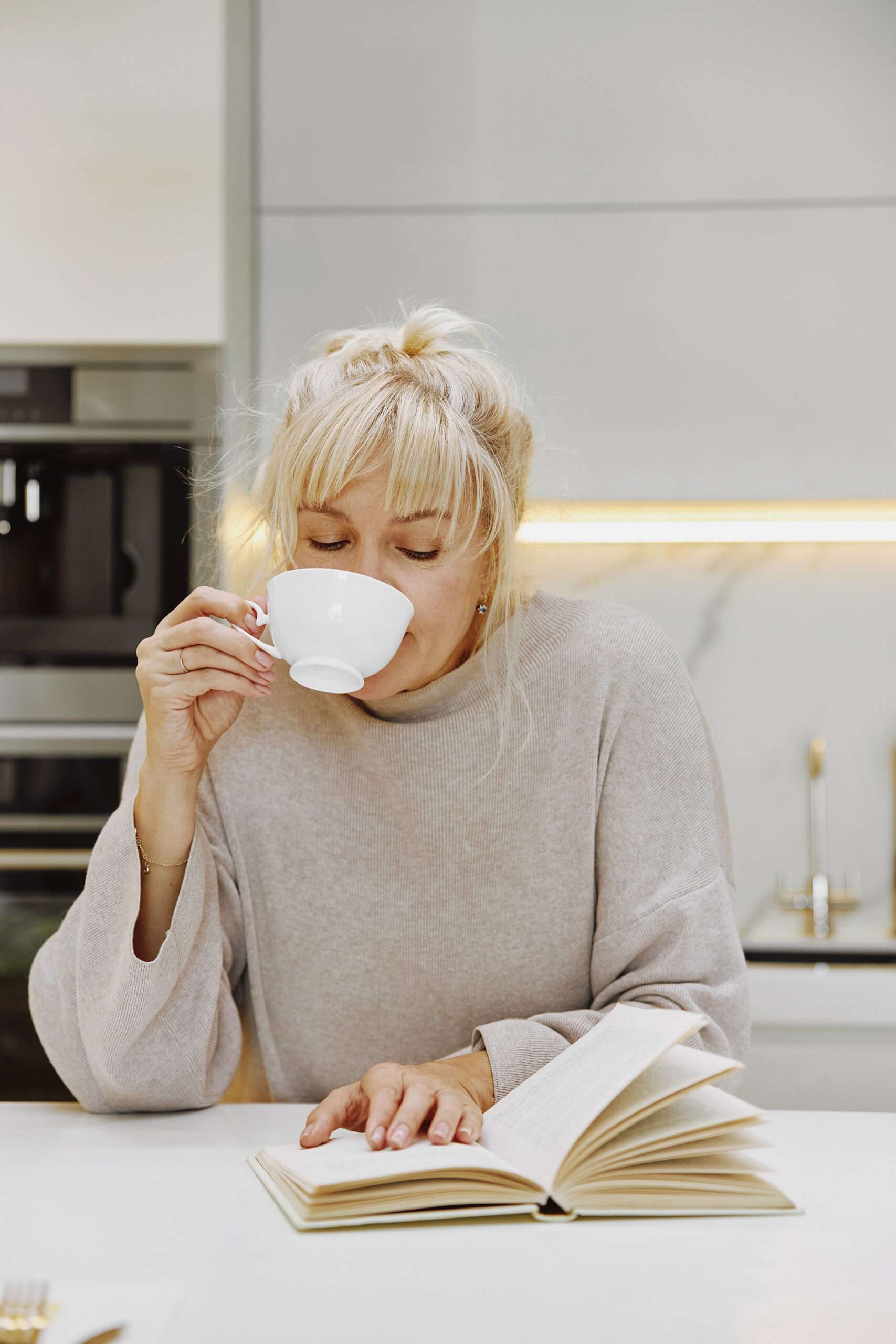 Woman leaning against a kitchen counter, drinking from a coffee cup while reading a book, appearing to multitask, representing the anxious feeling of the Sunday scaries.