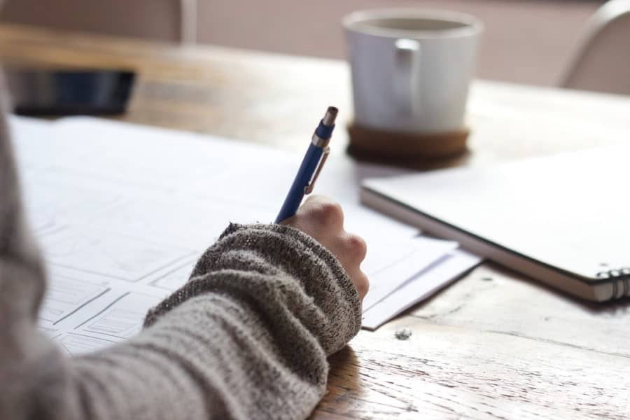 Woman’s right hand writing in a journal at a desk, viewed from behind, demonstrating the brain dumping process.