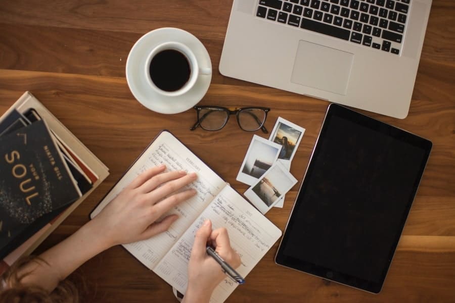 Overhead shot of a woman writing in a notebook on a desk with a cup of coffee, tablet, books, and laptop, symbolizing the organized benefits of brain dumping.