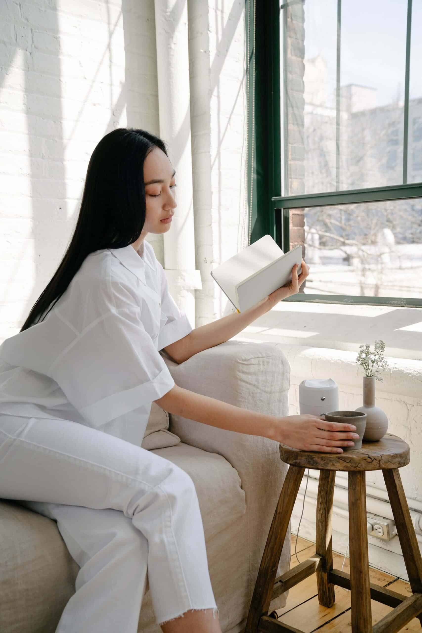 Woman sitting in an accent chair, holding a book and reaching for coffee on a side table, in a serene, minimalistic room, representing simple ways to care for yourself.