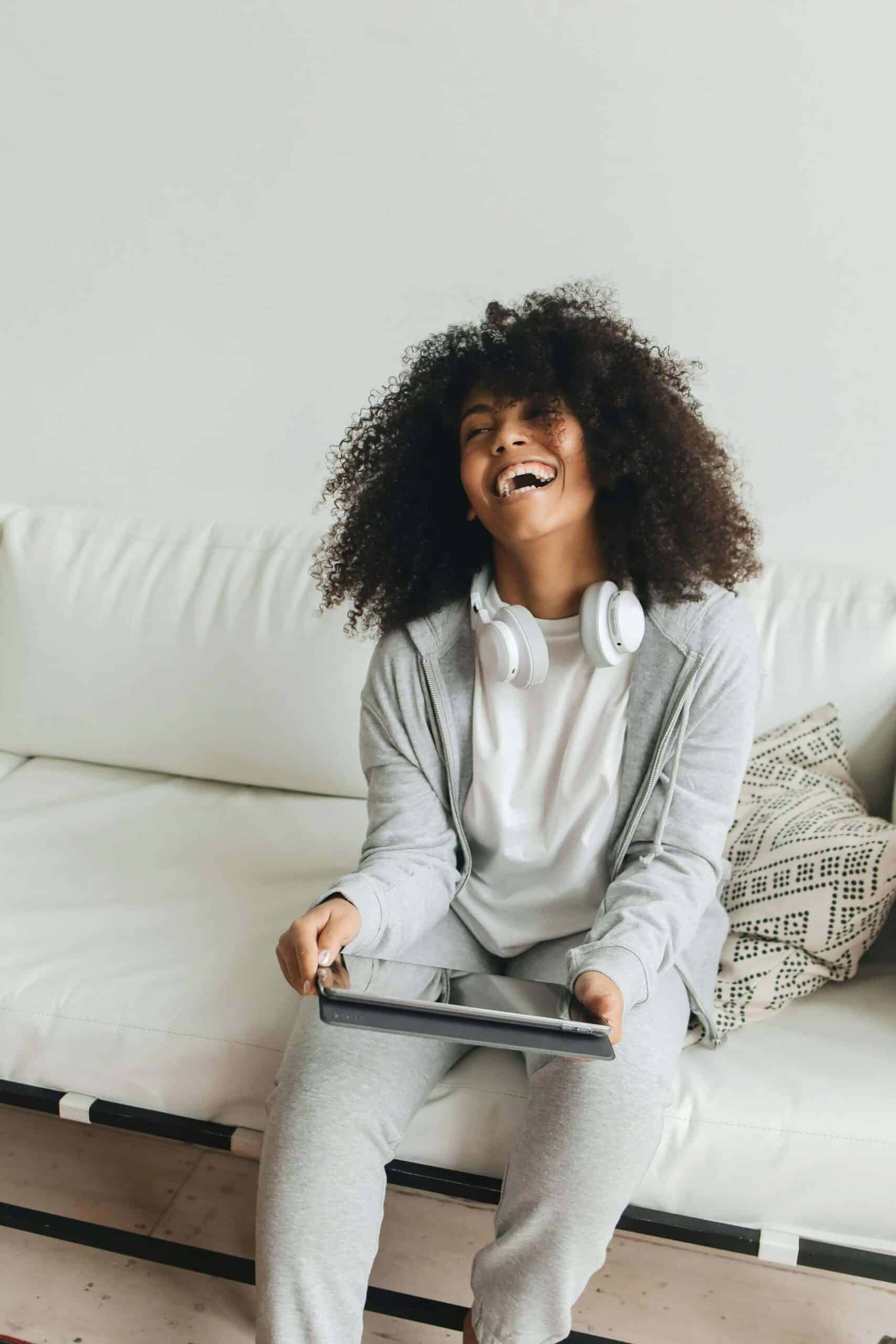 Woman sitting on a couch with a big smile, headphones around her neck, and a tablet in her hand, looking joyful and relaxed, showing how to improve your mood.