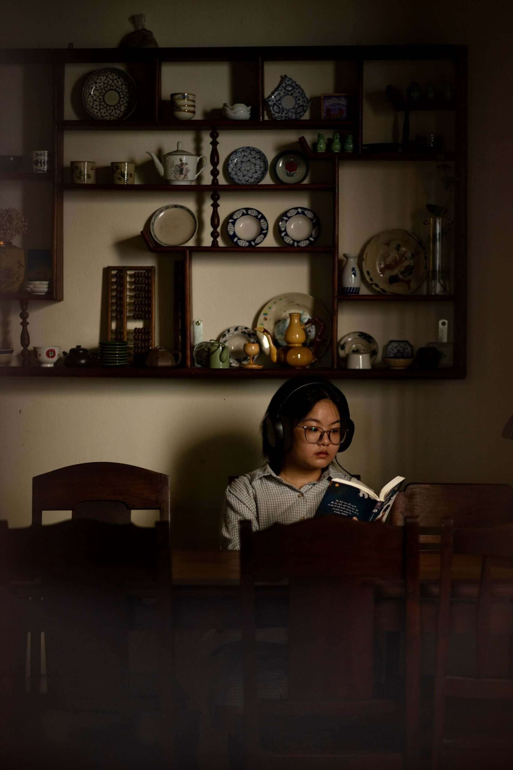 A woman sitting at a wooden table in a dimly lit room, reading a book while wearing headphones, fully engrossed, embodying a peaceful evening wind down.