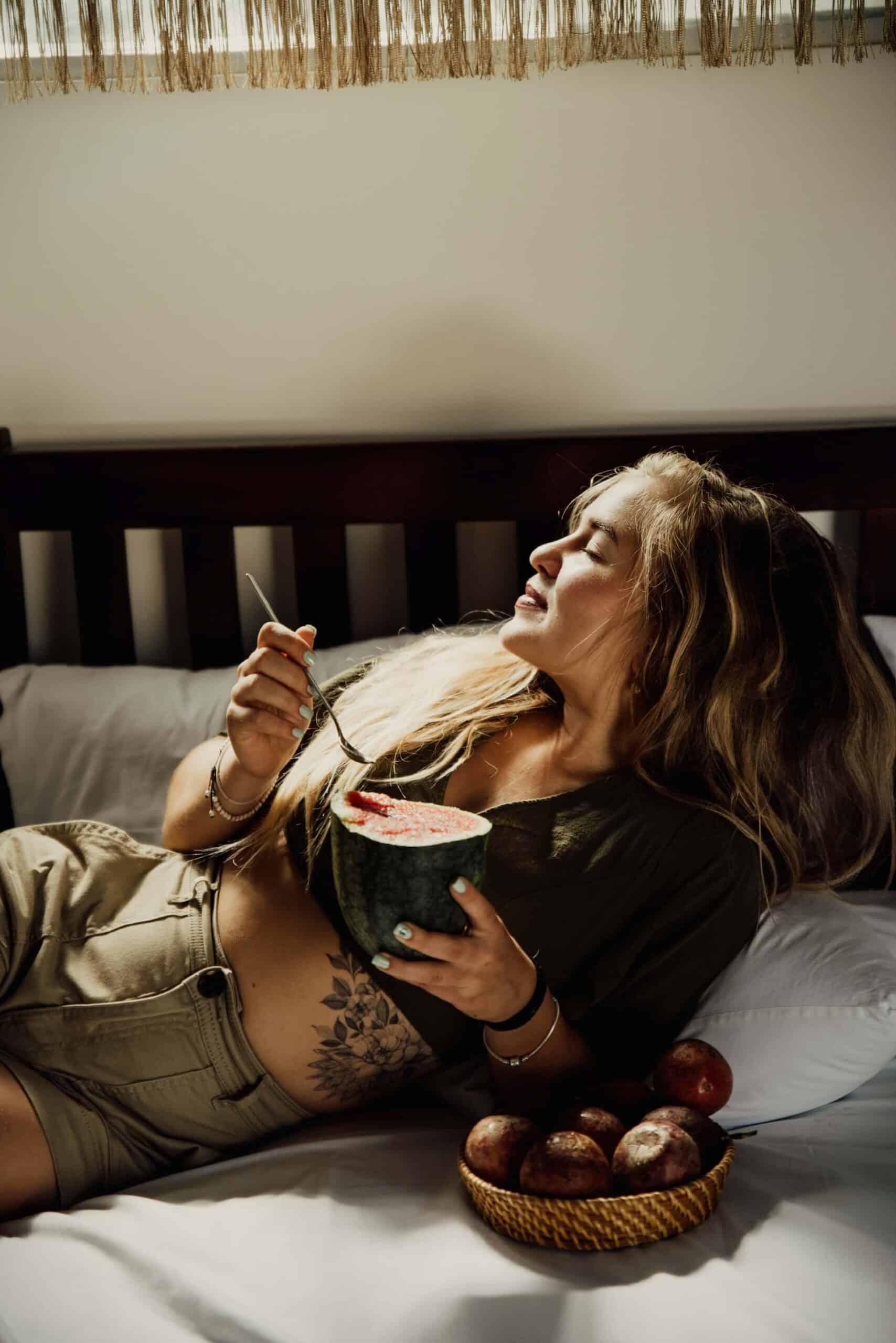 Woman leaning back on a bed, softly smiling while enjoying a half watermelon, exemplifying simple things to be grateful for, like savoring fresh fruit and relaxing moments.