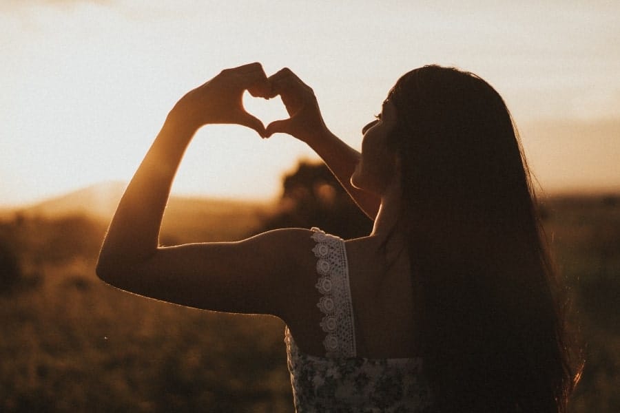 Woman facing a sunrise with her back to the camera, making a heart shape with her hands, symbolizing gratitude for a new day and the beauty of nature.