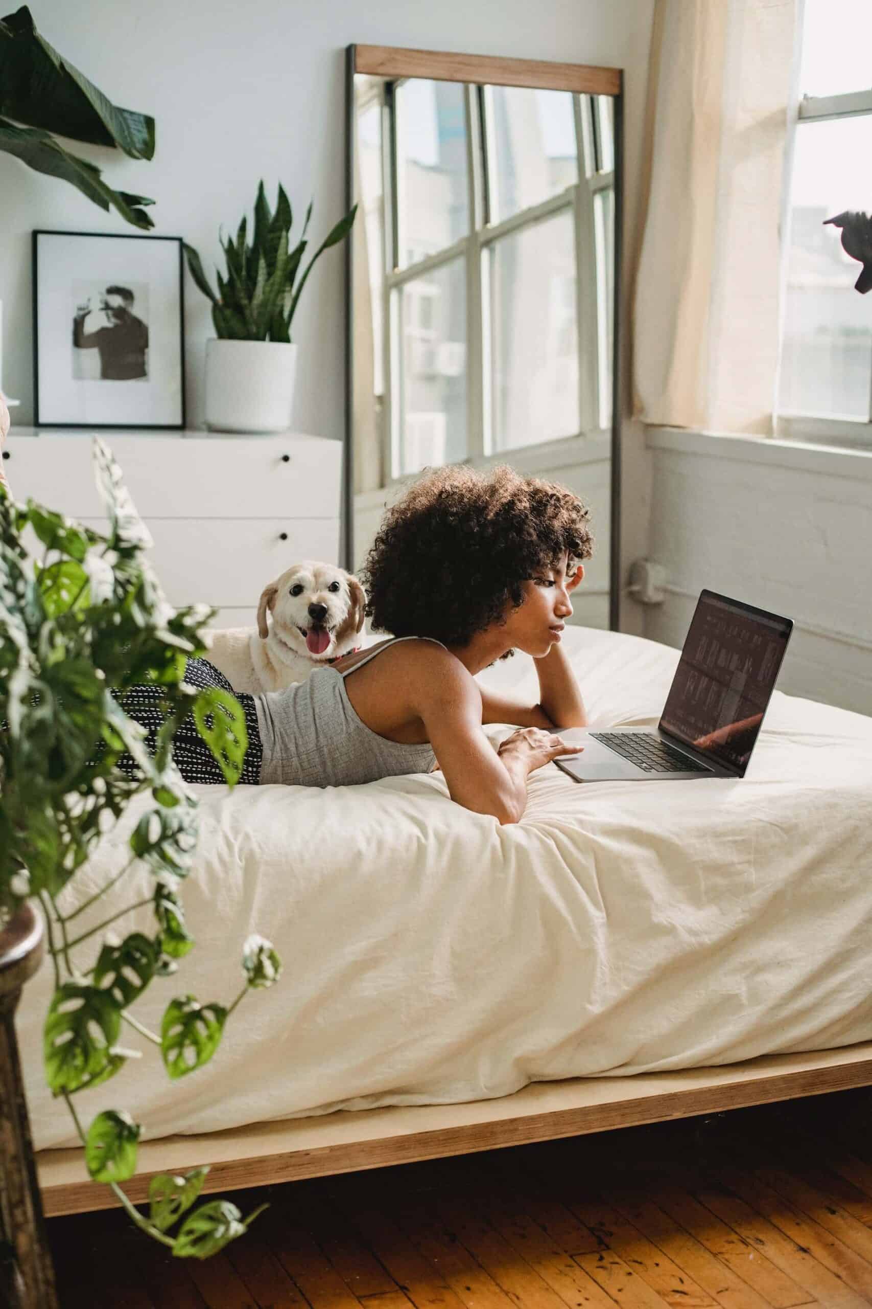 Woman lying on her stomach with a laptop in front of her while her dog rests its head on her back, contemplating mindset shifts.