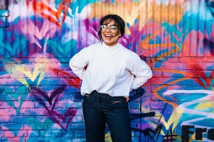 Woman smiling brightly in front of a colorful wall, reflecting gratitude for the creativity and vibrancy in the world around her.