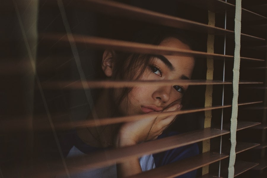 Woman looking out through window blinds, her concerned face partially turned towards the camera.