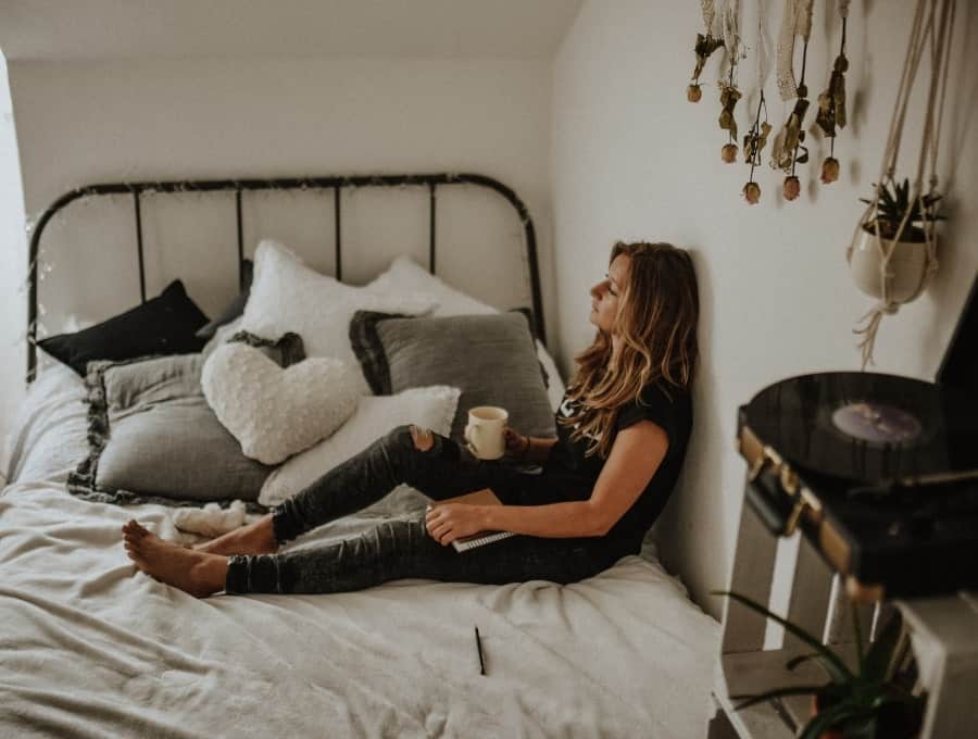 Woman sitting on a made bed with her back against the wall, holding a mug on her lap and gazing off, appearing to daydream.