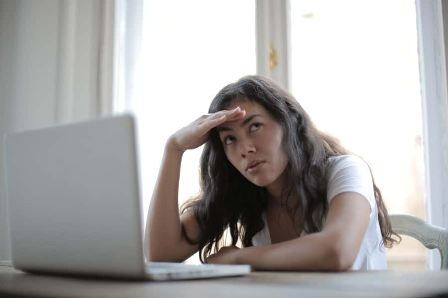 Woman sitting at a desk with an open laptop, her hand resting on her forehead as she gazes upward, indicating stress.