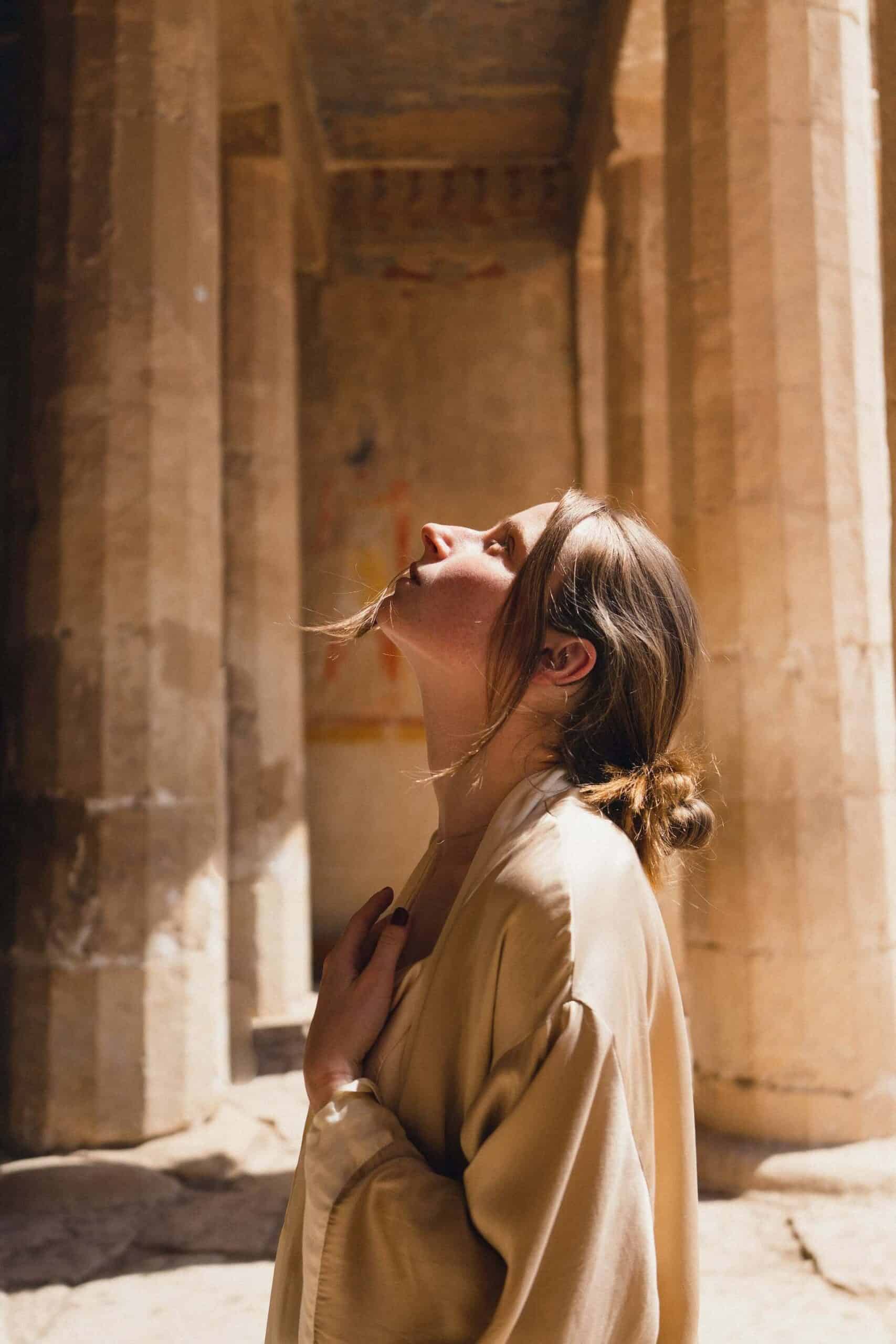 A side-profile view of a pensive woman standing in front of columns, looking up at the sky with her left hand resting on her chest, symbolizing introspection about a challenging job you hate.