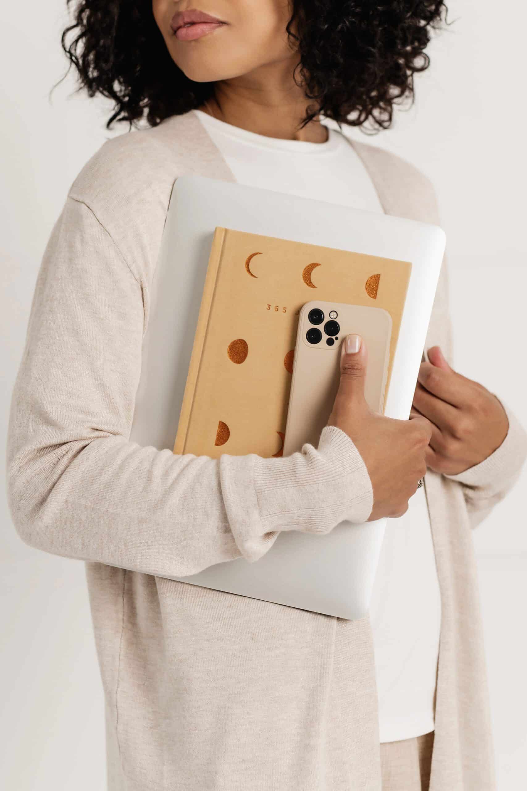 Woman holding a stack of items—a closed laptop, a journal, and a phone—against her chest, symbolizing organization and the intention to start fresh in the New Year.