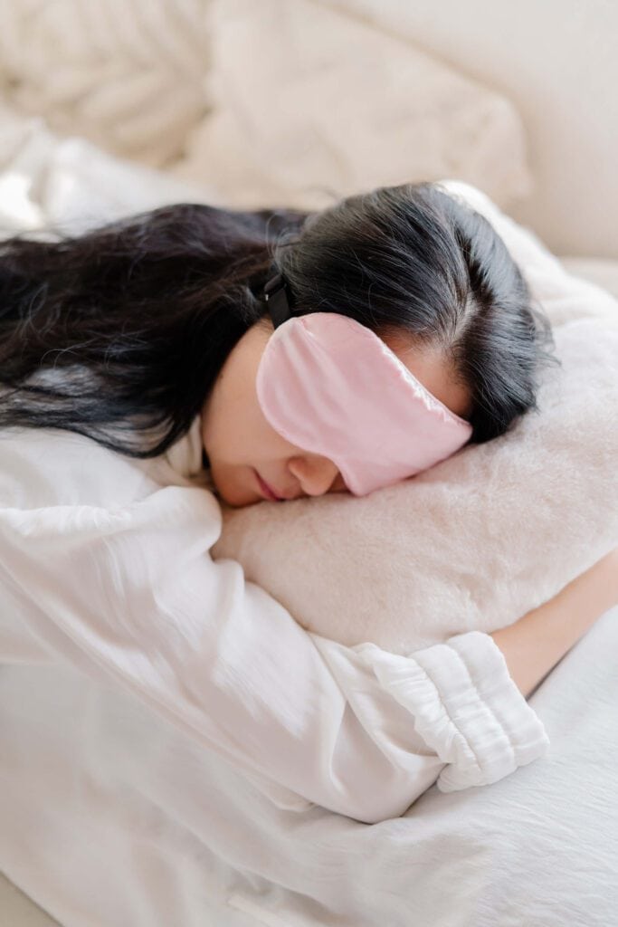 Woman sleeping on her stomach with a pink eye mask and light pink pajamas, illustrating the importance of quality sleep.