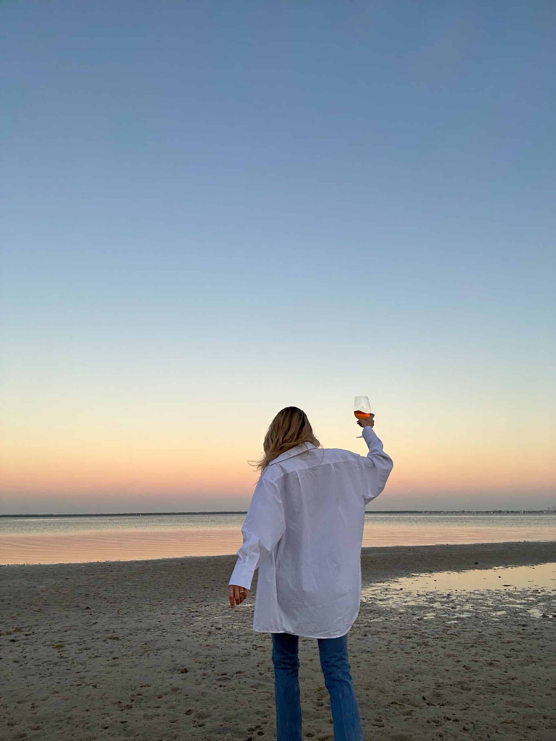 Woman in an open white button-down shirt, holding a glass of wine while swaying, representing the benefits of self-care and joyful relaxation.