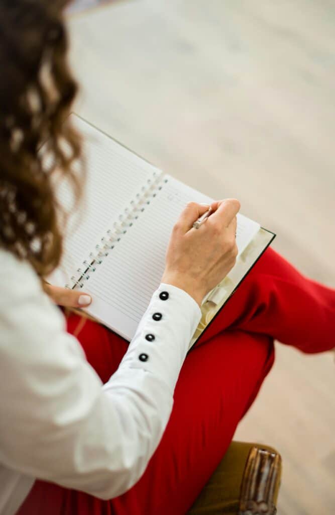 Over-the-shoulder view of a woman sitting cross-legged, writing in a notebook balanced on her knee, emphasizing goal-setting.